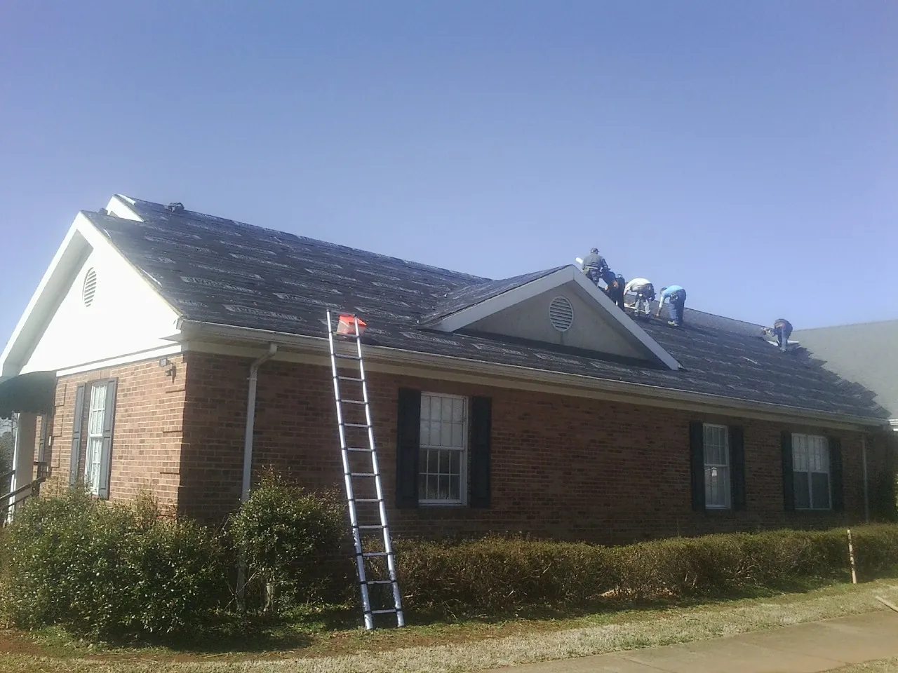Crew tearing shingles off a brick ranch roof with a ladder set — Emergency Roof Repair work by our crew in Cocolalla
