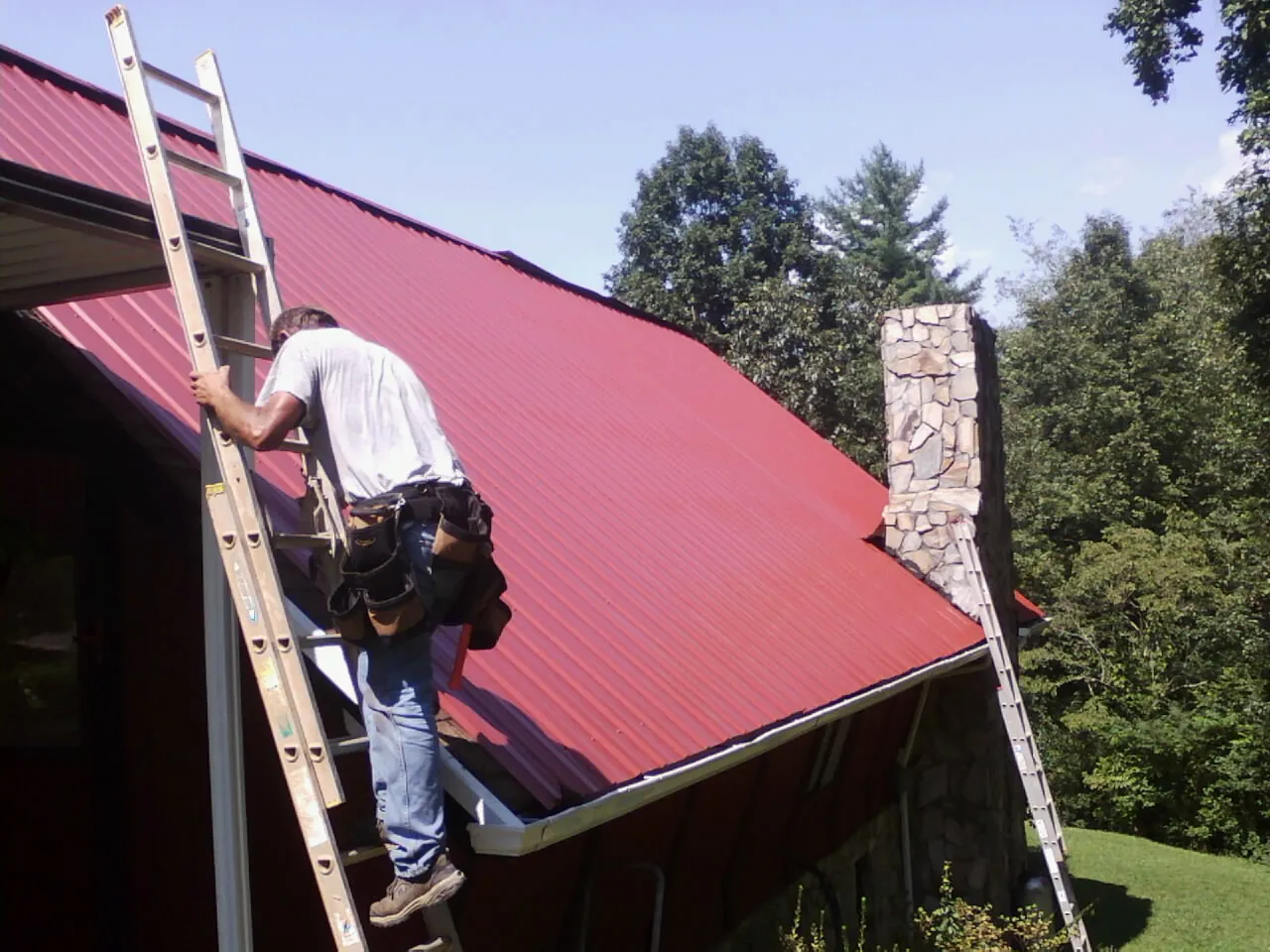Roofer on a ladder beside a red metal roof and stone chimney — Soffit & Fascia Repair by Roofing Brothers in Cocolalla